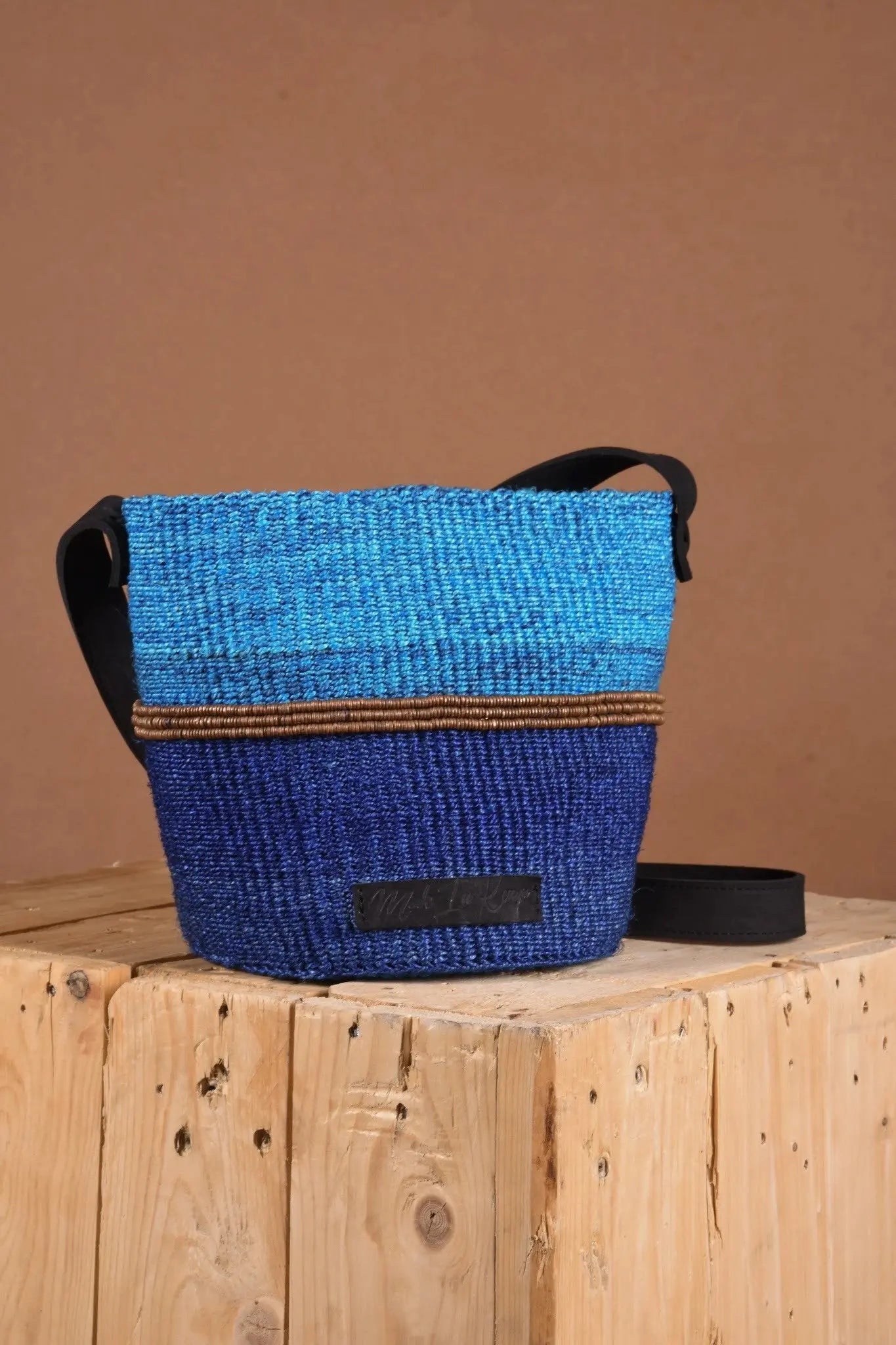 Blue woven bag with black accents on a wooden surface against a brown background. The bag is made from sustainably sourced sisal fibers