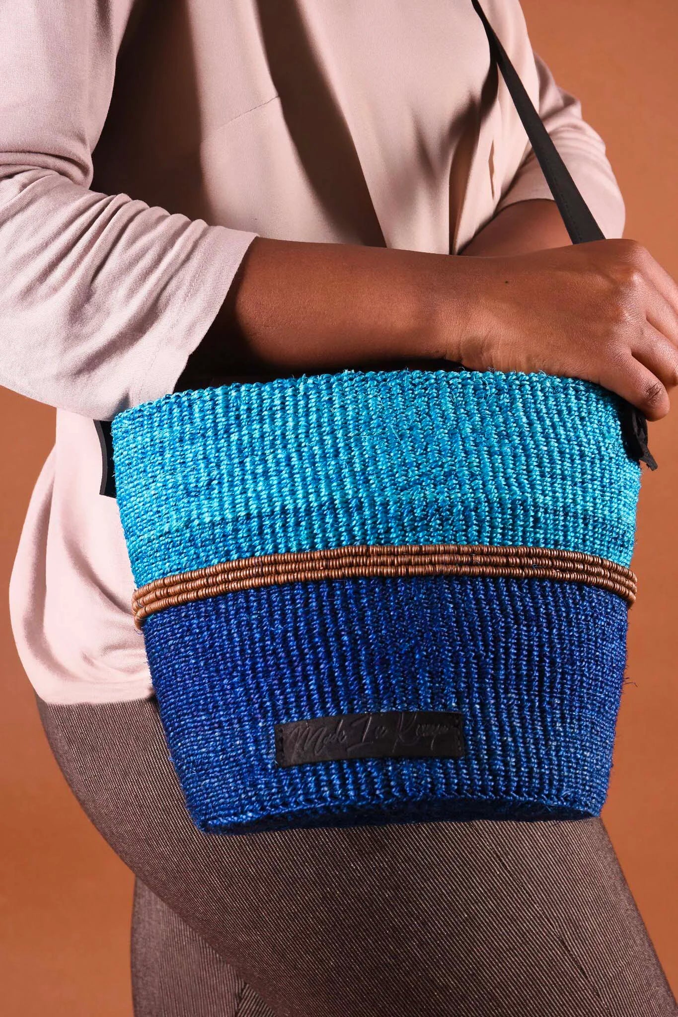 Person holding a blue woven bag with a brown stripe against a brown background. The bag is artisan-made from Africa