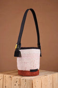Woven handbag with black strap on a wooden surface against a brown background. The bag is made by an all-women initiative using natural fibers