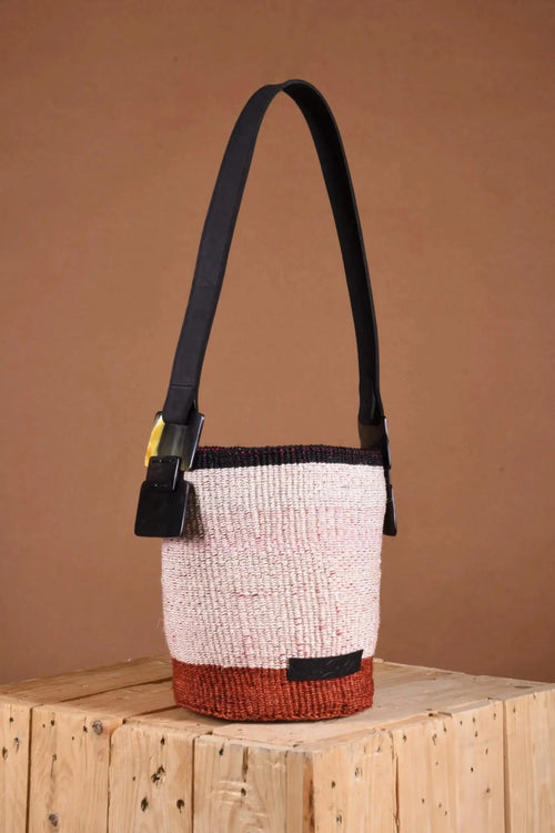 Woven handbag with black strap on a wooden surface against a brown background. The bag is made by an all-women initiative using natural fibers