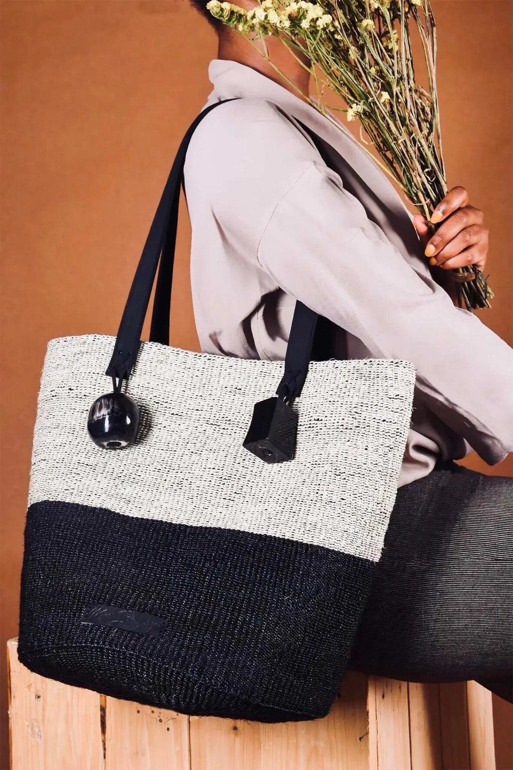 Person holding a black and white tote bag with flowers against a brown background. The bag is made from natural fibers by an all-women weaving initiative from Kenya-Africa