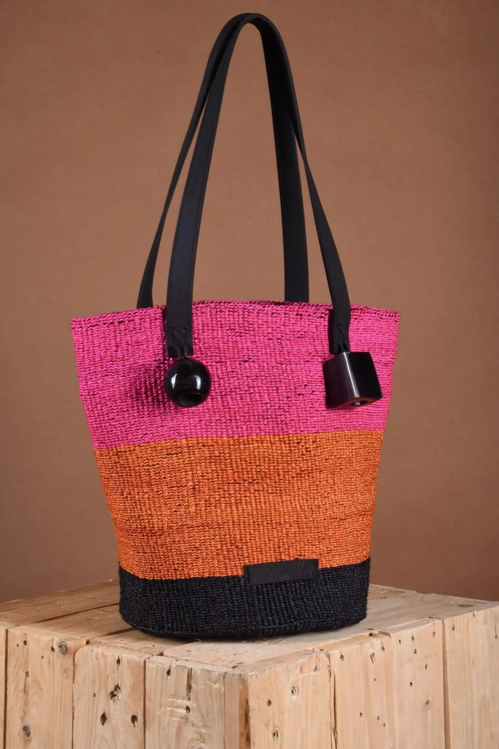 Tote bag with pink, orange, and black design on a wooden surface with a brown background. Women artisans make the bag using natural fibers and leather