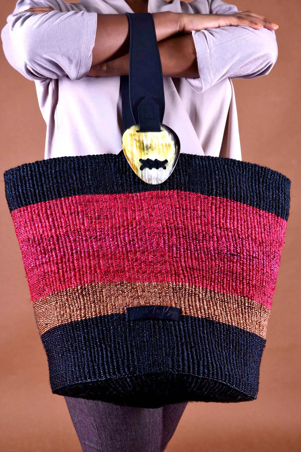 Multicolored striped bag with a decorative heart clasp held by a person against a brown background, the bag is made from sustainable materials by a women's weaving initiative from Kenya