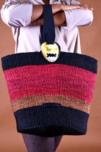 Multicolored striped bag with a decorative heart clasp held by a person against a brown background, the bag is made from sustainable materials by a women's weaving initiative from Kenya