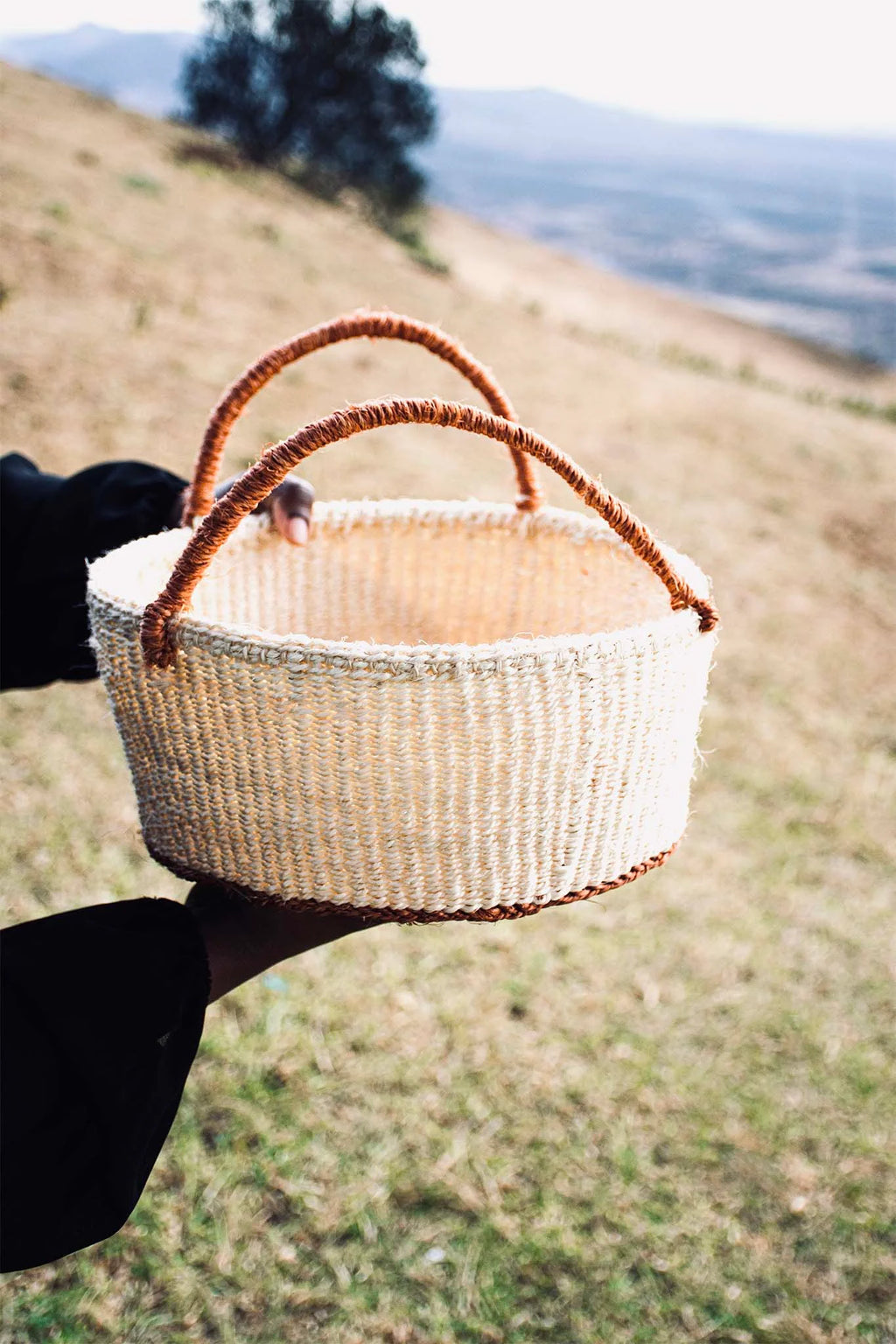 Person holding a woven basket in an outdoor setting with a blurred
landscape. Afrogaze basket