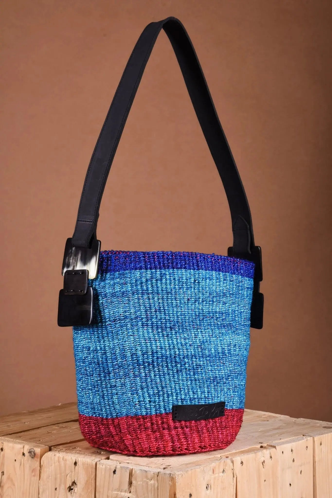 Blue and red woven bag with a black strap on a wooden surface and a brown background, made from natural fibers and leather by Women artisans