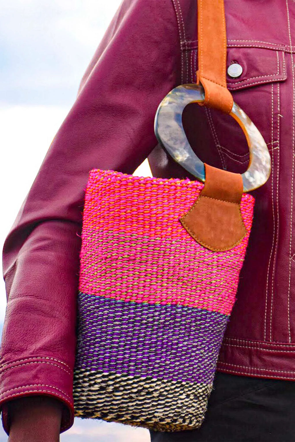 Colorful woven clutch bag with a leather strap held by a person wearing a maroon jacket. The bag is from afrogaze.com, and it's artisan-made